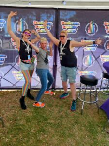 Dev, Tammi, and Zak have their arms up in excitement, posing in front of the Henderson Equality wall