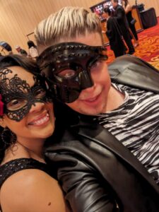 Crystal and Zak are dressed in black and wearing fancy black phantom of the opera masks. They are smiling. Zak has a zeebra print shirt on.