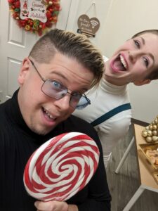 Zak looks happy holding a huge red and white swirl lollipop. Dev laughs in the background.