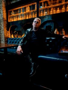 A very dark photo with gold accents as Zak sits at a bar, leather back seating, he is dressed in black with knee high lace up boots. He looks serious and fashionable.