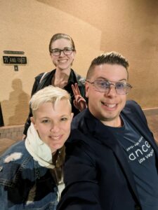 Tammi, Dev, and Zak pose in front of a restaurant wall, casually dressed.